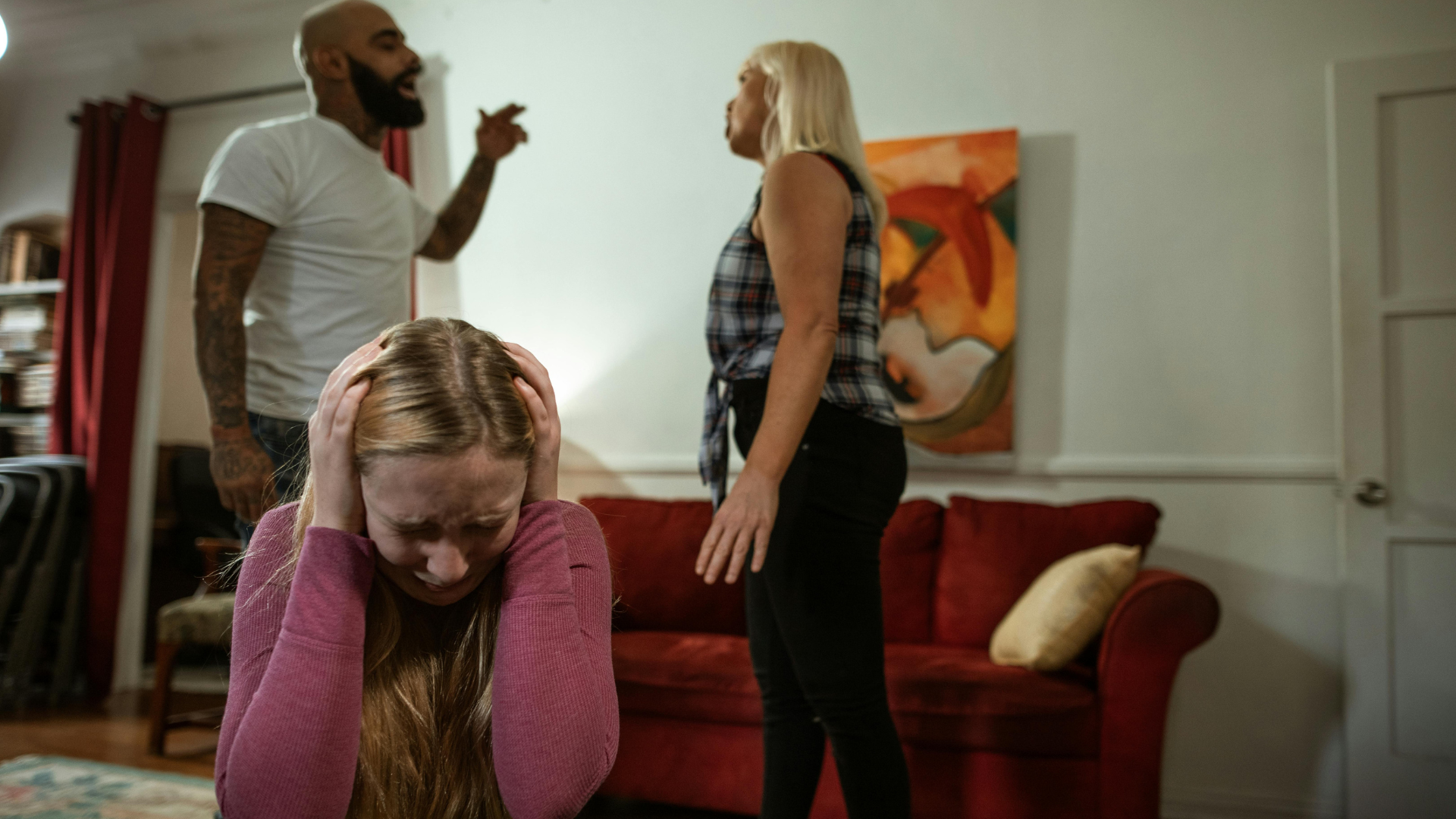 Terapia Integrativa, a imagem mostra uma cena de três pessoas em um ambiente interno. No centro, uma mulher loira está sentada em um sofá vermelho, com as mãos na cabeça, parecendo angustiada. Atrás dela, um homem careca com barba e tatuagens, vestindo uma camiseta branca, está apontando e parece estar gritando. À direita, uma mulher loira de pé, vestindo uma camisa xadrez, está olhando para o homem. A expressão dela não é clara, mas parece estar envolvida na situação. Ao fundo, há uma estante com livros e uma cortina vermelha.